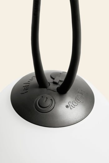 Close-up of the Fatboy Bolleke Mini La Surprise by Fatboy: a black circular base with embossed buttons and power symbol, two black cords extending upward, and adjustable dimming. The spherical lamp sits on a smooth white surface.