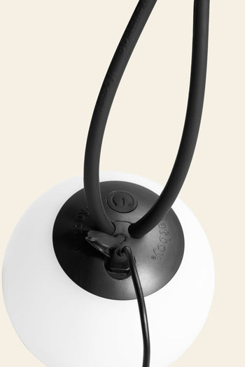 Close-up of the Fatboy Bolleke Mini La Surprise by Fatboy—a black and white spherical lamp base with a power cable, two flexible black arms, and adjustable dimming, set against a plain light background.