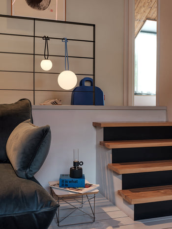 Modern living space with wooden stairs, a blue suitcase, and Fatboy Bolleke Mini La Surprise spherical lamps casting a warm glow. A small table holds a blue book and glass object by a dark sofa. Sneakers are visible behind the black metal railing.