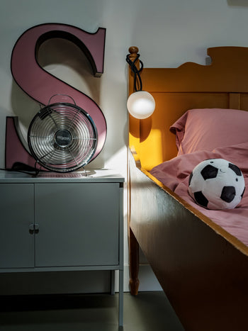 A bedside scene featuring the Fatboy Bolleke Mini La Surprise lamp, a large pink letter S, a metal fan, and a white cabinet next to a yellow bed with pink bedding and a plush soccer ball. The lamp is mounted above the headboard.