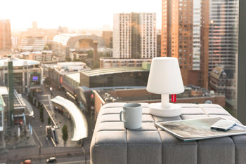 A Fatboy Edison the Petit lamp, a gray mug, a magazine, and a smartphone sit on an ottoman by a large window with sunset views of tall city buildings.