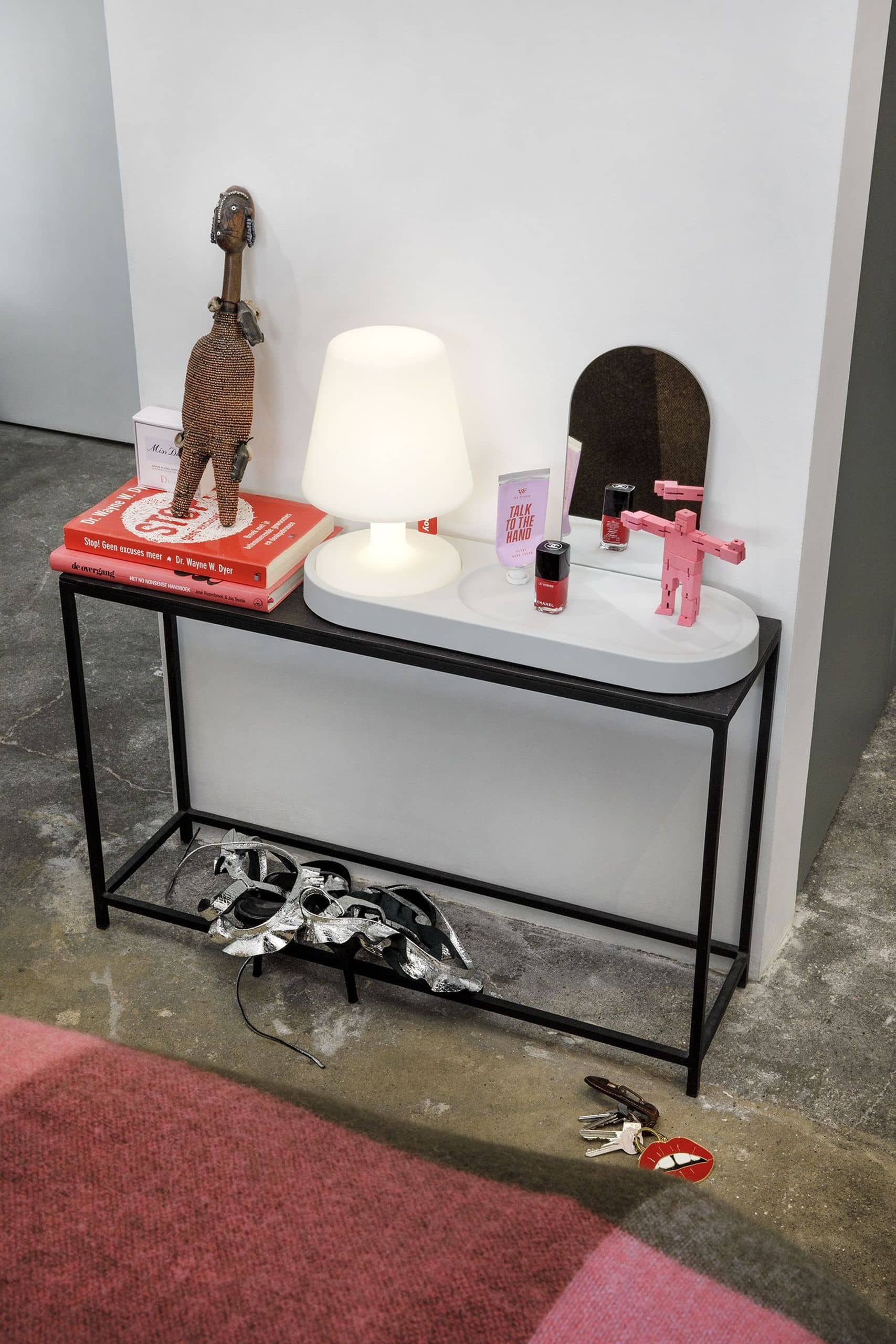 A small table against a white wall holds the Fatboy Edison the Petit lamp, sculpted figures, books, a small round mirror, and nail polish. Below, a silver mask sits on the lower shelf with keys and a keychain on the floor nearby.