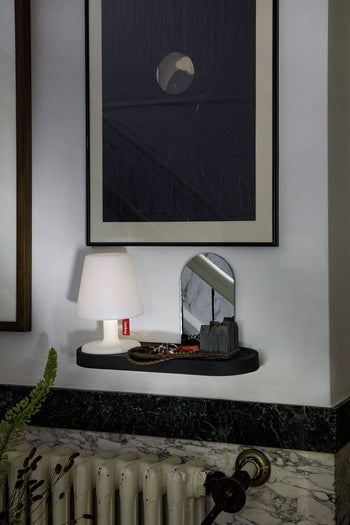 A Fatboy Edison the Petit lamp, a mirror, and assorted items sit on a black marble shelf under a framed abstract print. The arrangement is above a radiator with diffused light and a plant partially seen in the foreground.