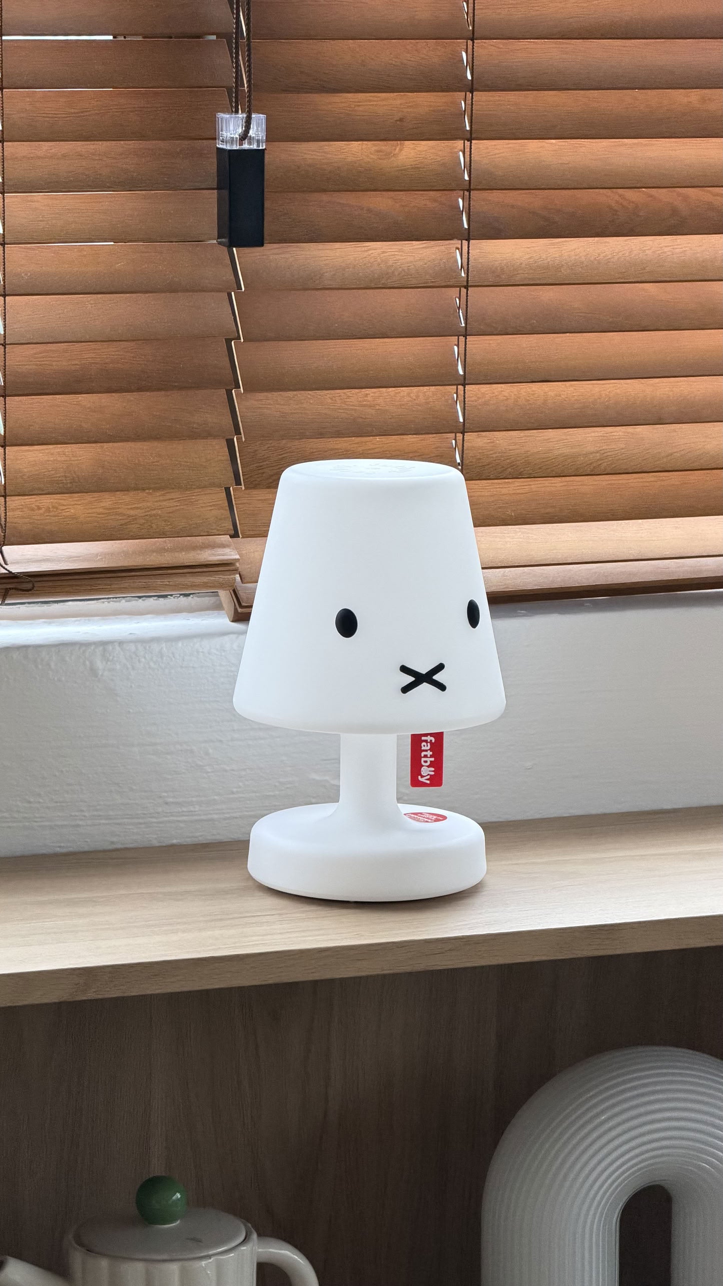 The Fatboy Edison the Petit x Miffy lamp by Fatboy, shaped like Miffy’s cartoon rabbit face, sits on a shelf. This portable and rechargeable LED lamp is perfect as a table light for any space.