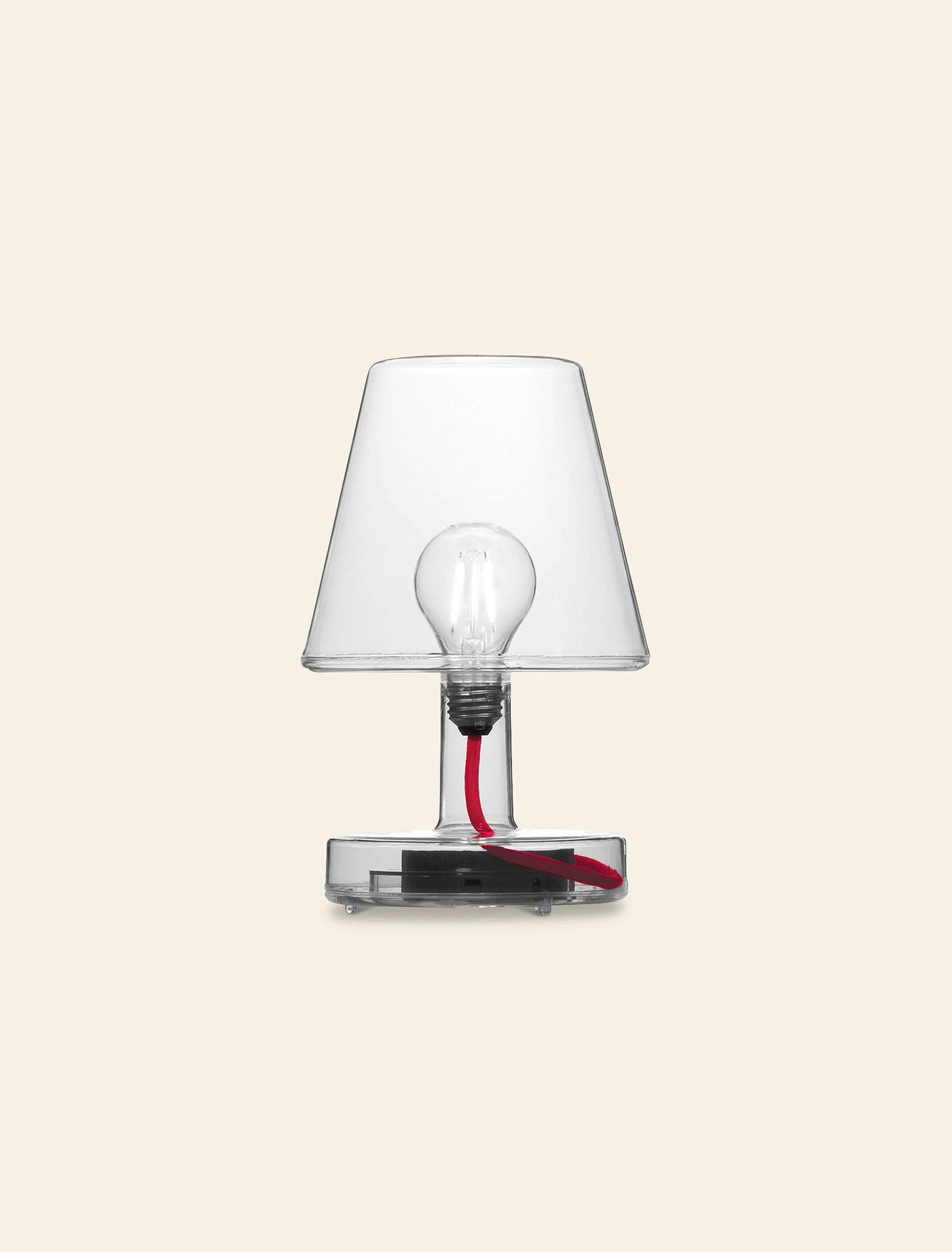 The Fatboy Transloetje by Fatboy is a modern table lamp with a transparent base and shade, visible bulb, and red cord—an innovative lighting piece that adds unique style to any room, shown here on a plain beige background.