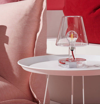 The Fatboy Transloetje clear glass lamp by Fatboy, featuring a visible bulb, sits on a white round side table beside a pink pillow and red cushion in a bright, modern room, highlighting its innovative and stylish design.
