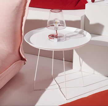 A modern white round side table with thin metal legs displays the Fatboy Transloetje lamp, adding unique lighting by Fatboy. Soft red and pink cushions and white geometric furniture complete the brightly lit scene.