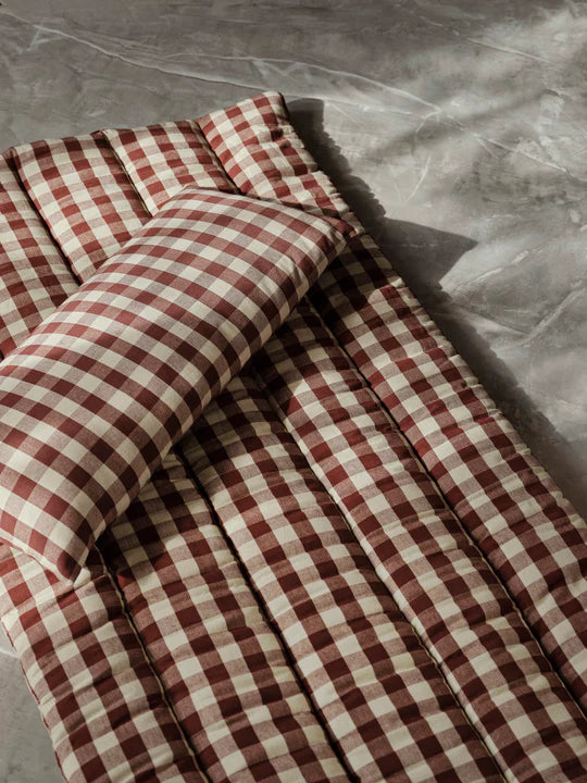 The Ferm Living Bothy Check Quilted Mattress, paired with a matching pillow in red and white checks, rests on a gray textured surface as sunlight highlights this rustic decor from Ferm Living.