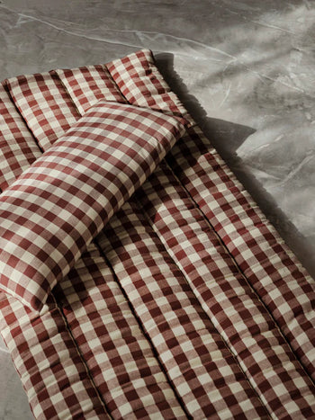 The Ferm Living Bothy Check Quilted Mattress, paired with a matching pillow in red and white checks, rests on a gray textured surface as sunlight highlights this rustic decor from Ferm Living.
