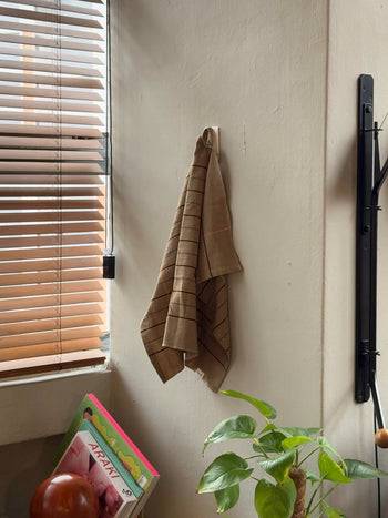 The Ferm Living Ito Tea Towel, Set of 2, hangs on a wall hook near a window with wooden blinds. Below, a green potted plant sits beside colorful magazines on the table as warm natural light brightens the cozy room.