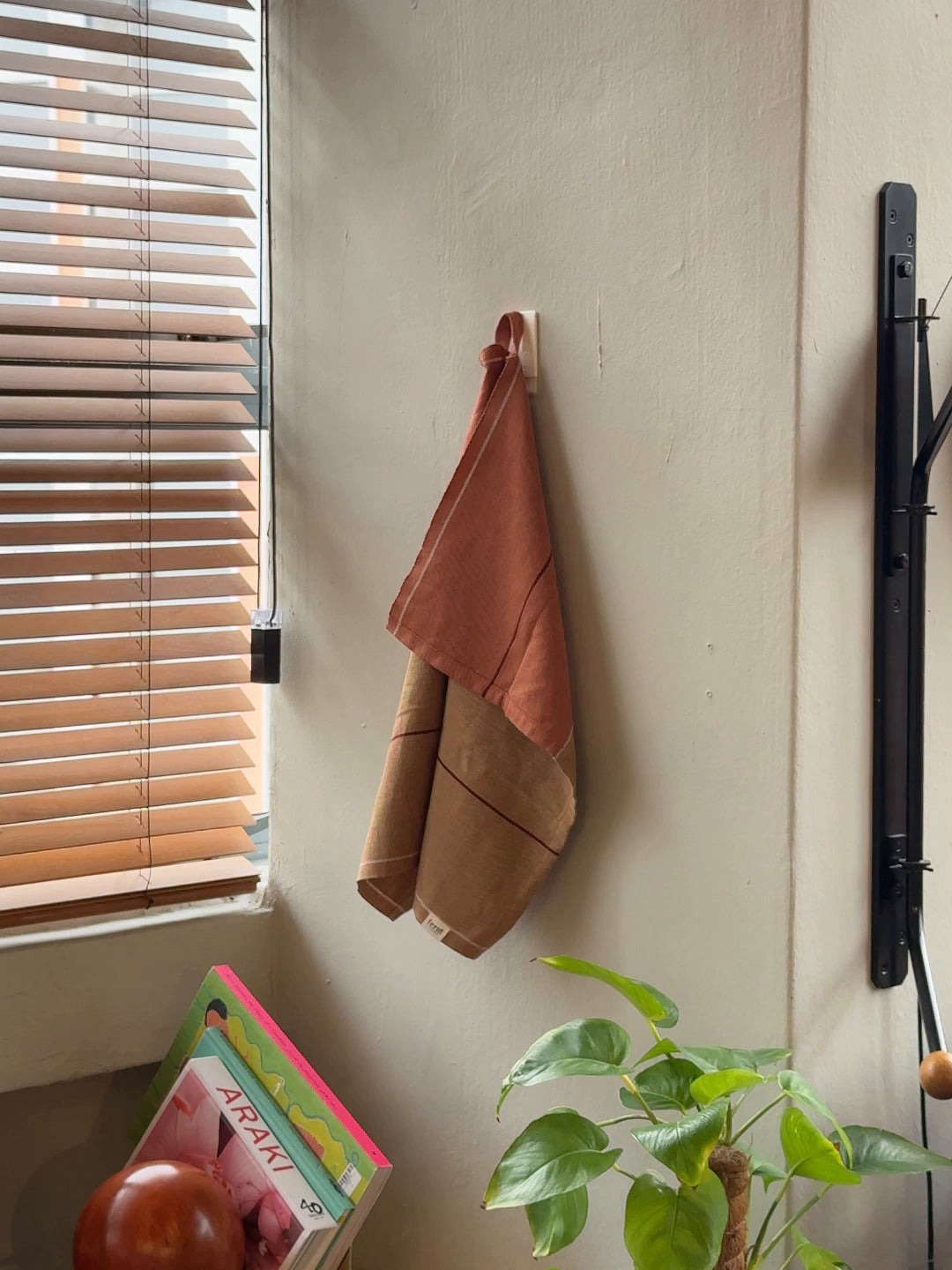 The Ferm Living Ito Tea Towel, Set of 2, in brown and rust organic cotton, hangs on a wall hook by a window with wooden blinds. A green plant and colorful books or magazines are stacked on the table below.