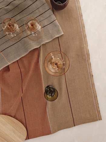 Overhead view of three empty wine glasses and one green glass on Ferm Living Ito Tea Towels (set of 2) with a striped, earth-toned twill weave. A wooden board sits in the corner and a bowl is partially visible at the top.