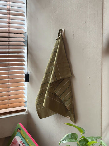 A set of two Ferm Living Ito Tea Towels in green check hangs on a light wall by a window with brown blinds, with a leafy plant and part of a colorful book visible below.