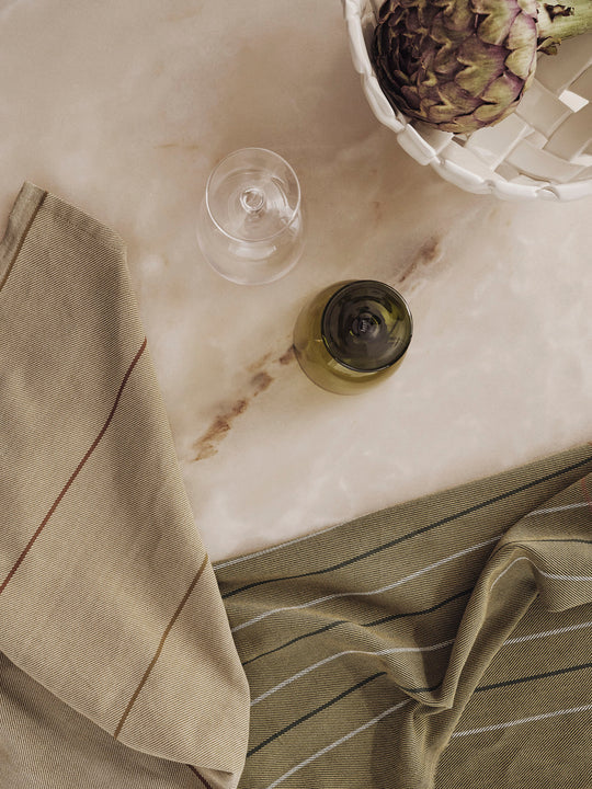 A top-down view of a beige marble surface with a wine glass, green bottle, and two striped Ferm Living Ito Tea Towels (set of 2) by Ferm Living, plus a white basket holding an artichoke.