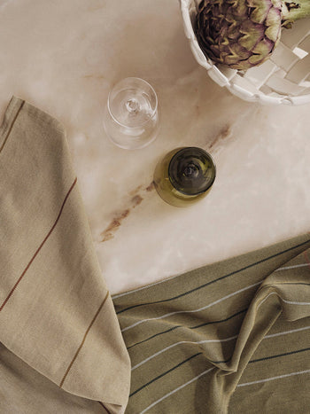 A top-down view of a beige marble surface with a wine glass, green bottle, and two striped Ferm Living Ito Tea Towels (set of 2) by Ferm Living, plus a white basket holding an artichoke.