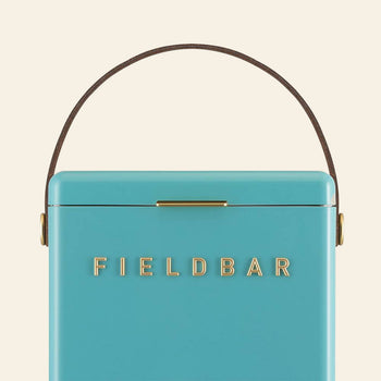 A close-up of the turquoise Fieldbar Drinks Box by Fieldbar, featuring a brown handle and gold FIELD BAR lettering on the front, set against an off-white background.