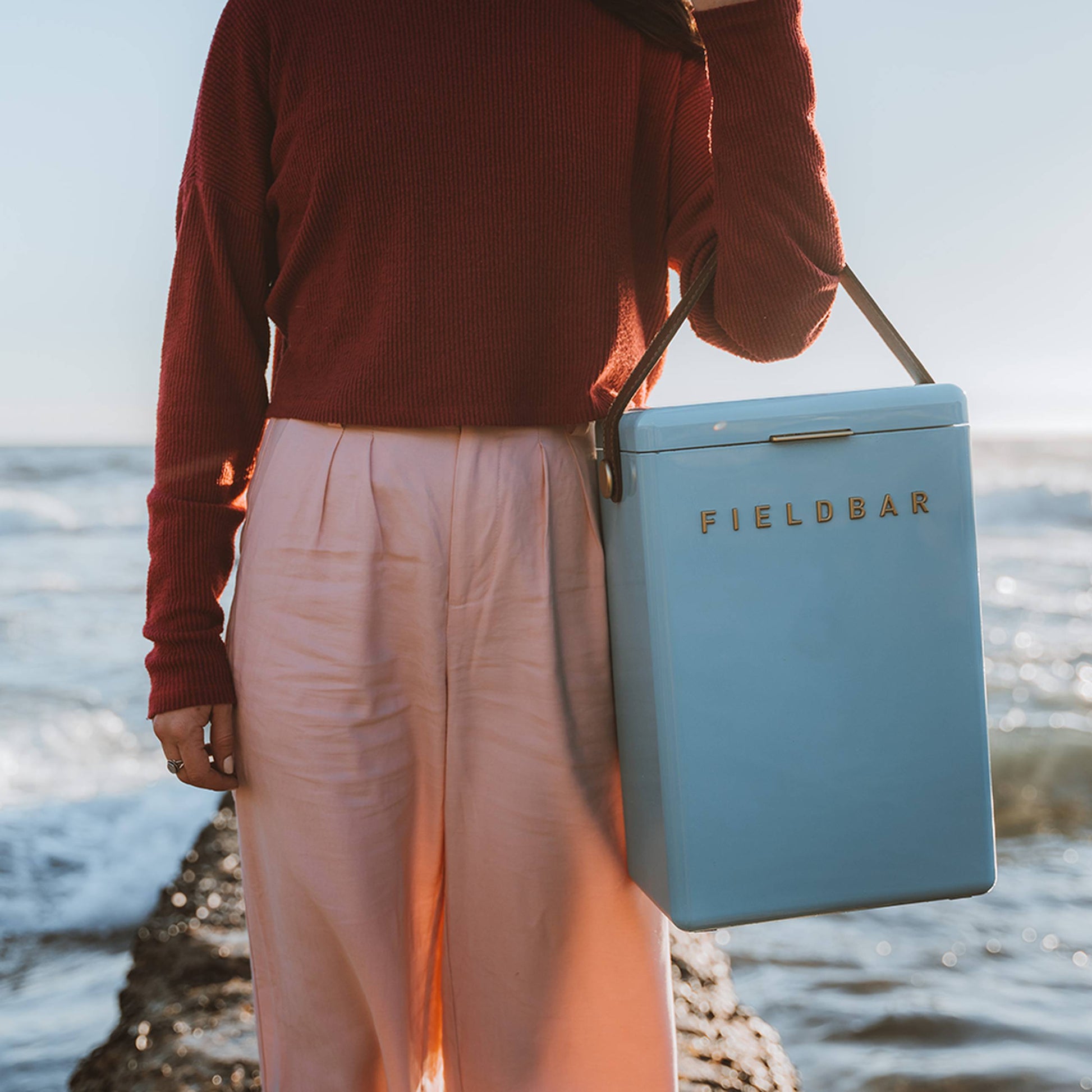By the sea, someone in a red long-sleeve top and pink pants carries a pastel blue Fieldbar Drinks Box by Fieldbar with a black strap. Waves and blue sky set the scene for this blend of style and thermal performance at the coast.