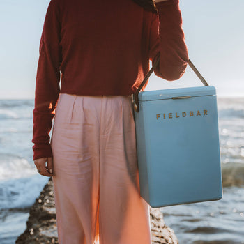 By the sea, someone in a red long-sleeve top and pink pants carries a pastel blue Fieldbar Drinks Box by Fieldbar with a black strap. Waves and blue sky set the scene for this blend of style and thermal performance at the coast.