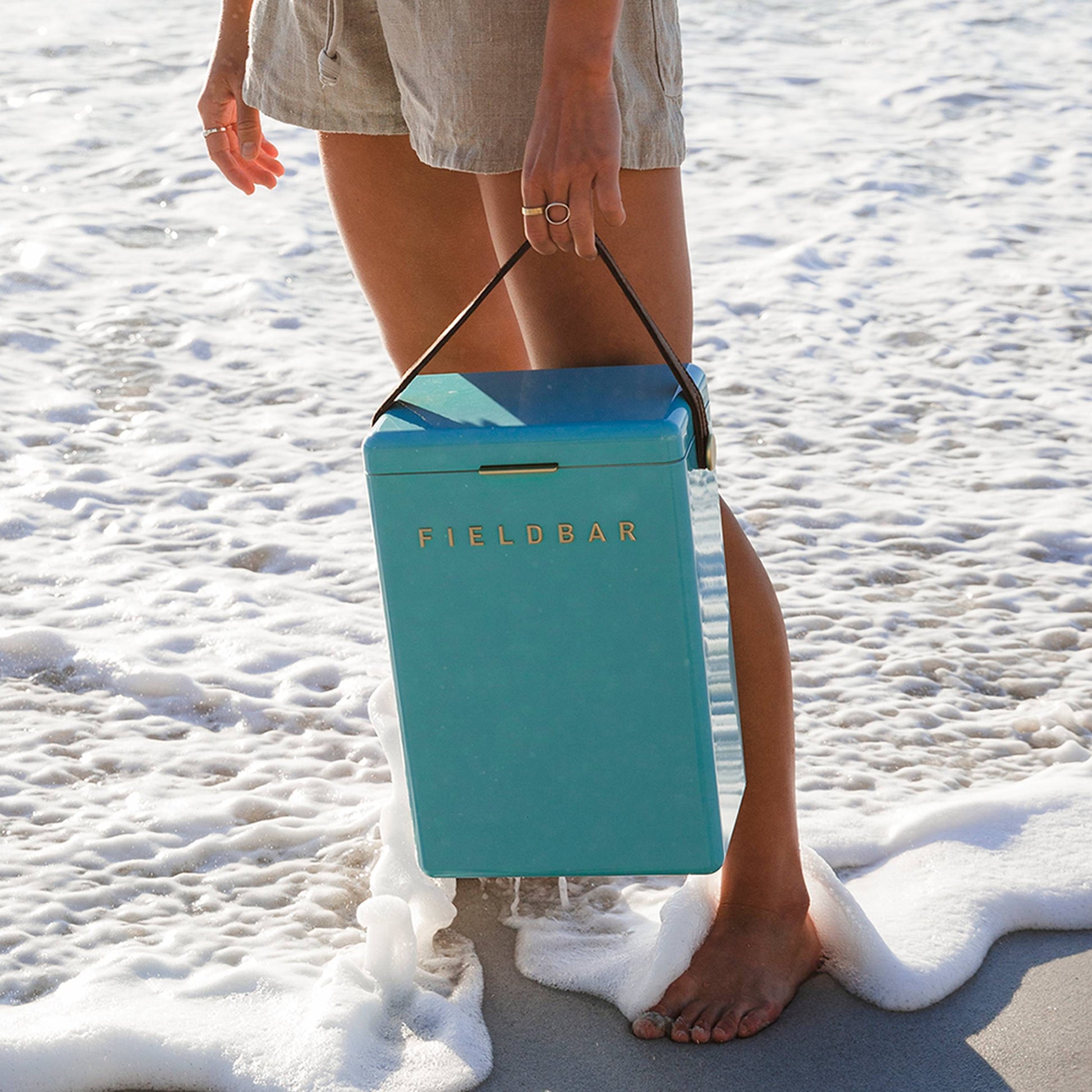 Standing barefoot on the sandy shore, a person holds a teal Fieldbar Drinks Box by its handle. Gentle waves lap their feet, emphasizing the Fieldbar coolers stylish design and outstanding thermal performance.