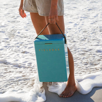 Standing barefoot on the sandy shore, a person holds a teal Fieldbar Drinks Box by its handle. Gentle waves lap their feet, emphasizing the Fieldbar coolers stylish design and outstanding thermal performance.
