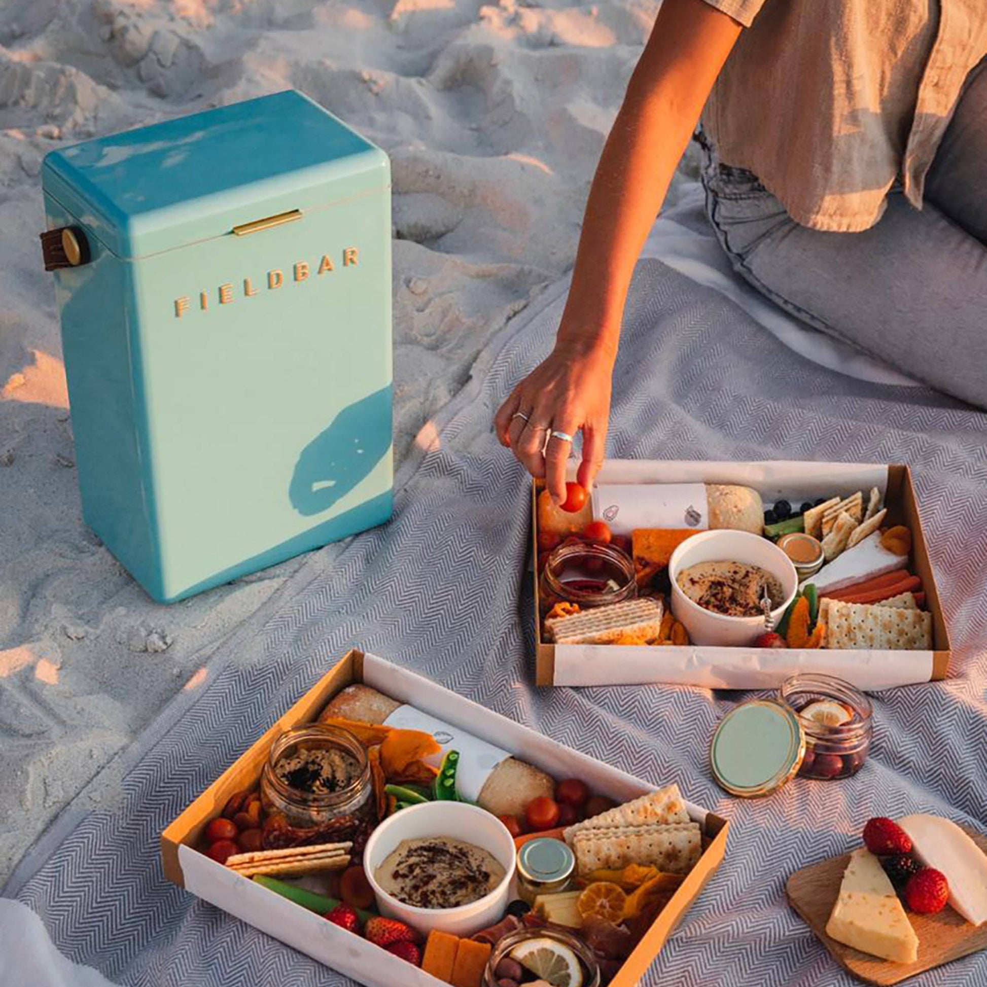 At the beach, someone sits on a blanket with two picnic snack boxes and a turquoise Fieldbar Drinks Box cooler from Fieldbar, which keeps fruits, cheese, crackers, dips, and other finger foods fresh thanks to its excellent thermal performance.