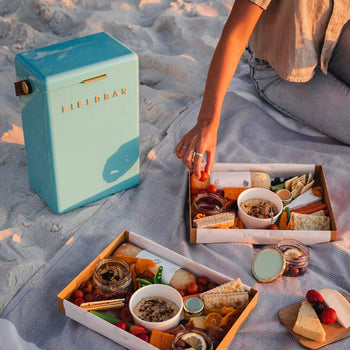 At the beach, someone sits on a blanket with two picnic snack boxes and a turquoise Fieldbar Drinks Box cooler from Fieldbar, which keeps fruits, cheese, crackers, dips, and other finger foods fresh thanks to its excellent thermal performance.