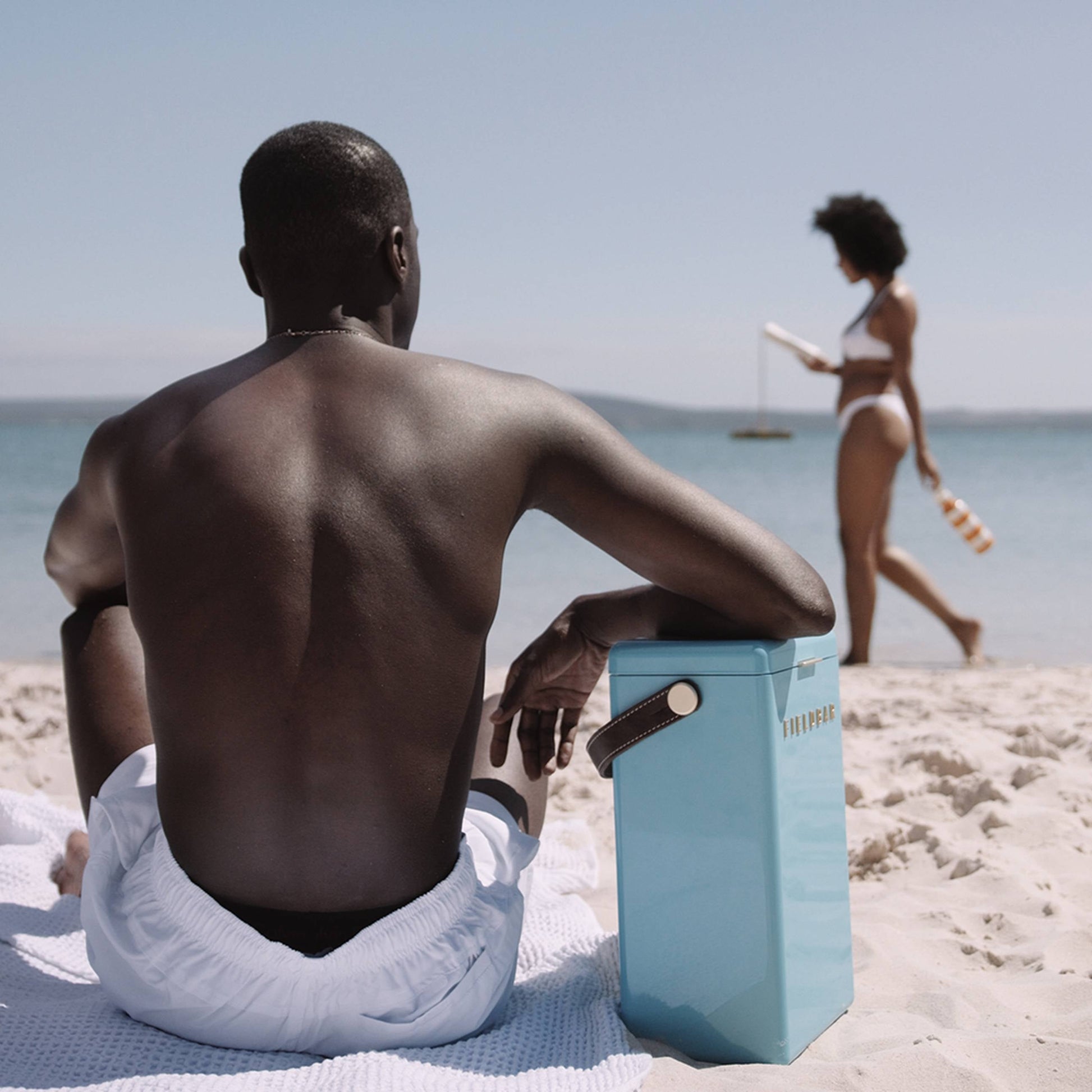 A man in white shorts sits on a sandy beach facing the sea, with a blue Fieldbar Drinks Box by Fieldbar beside him. In the background, a woman in a bikini walks along the shore holding a bottle under a clear sky.