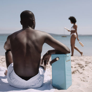 A man in white shorts sits on a sandy beach facing the sea, with a blue Fieldbar Drinks Box by Fieldbar beside him. In the background, a woman in a bikini walks along the shore holding a bottle under a clear sky.