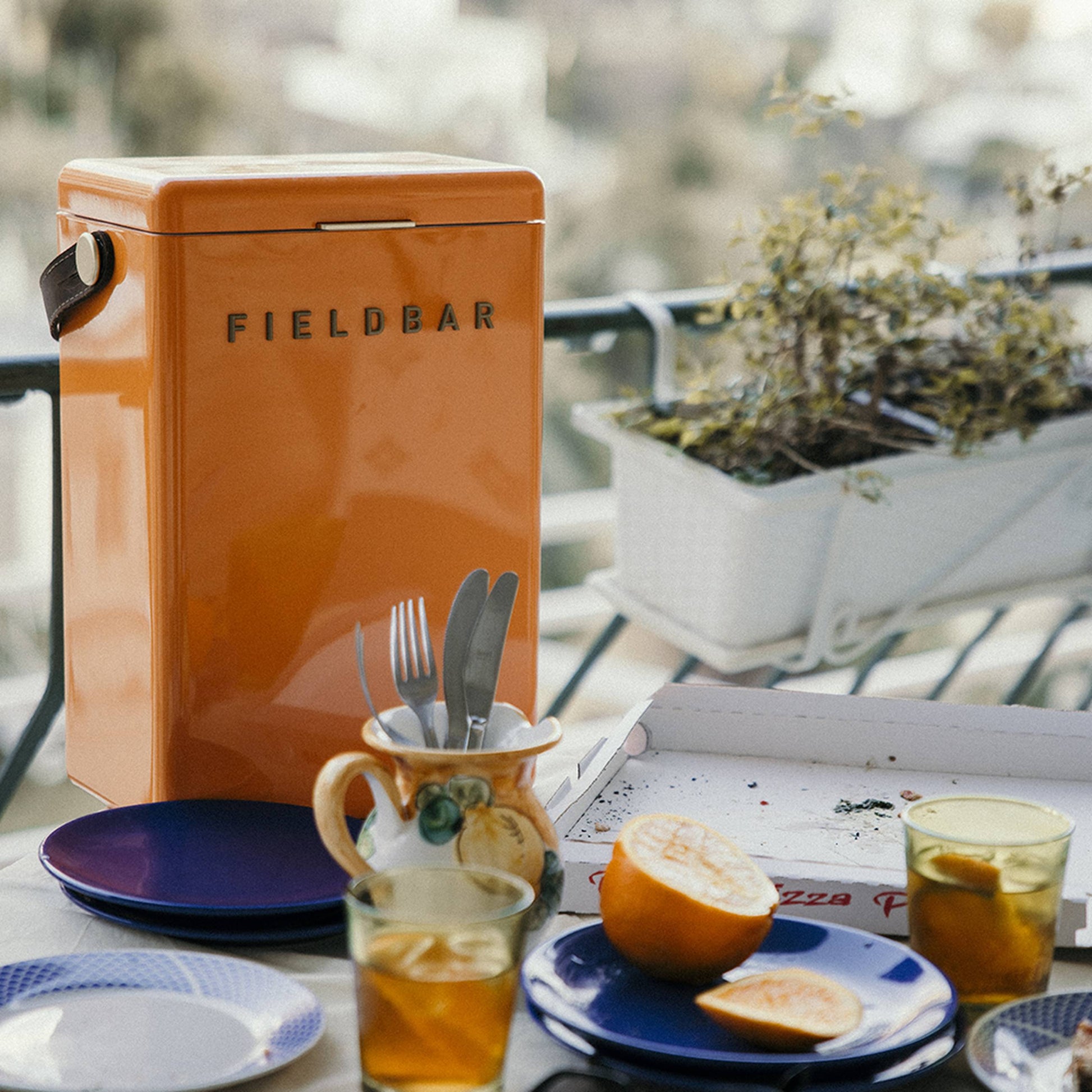 A Fieldbar Drinks Box by Fieldbar in orange sits on an outdoor table with plates, iced tea glasses, cutlery in a cup, and a partly eaten pizza. A planter filled with greenery hangs from the railing behind the table.