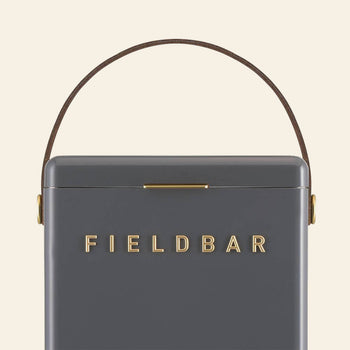 A close-up of the sleek, dark gray Fieldbar Drinks Box by Fieldbar features gold branding on the front, a slim brown side handle, and highlights its exceptional thermal performance.