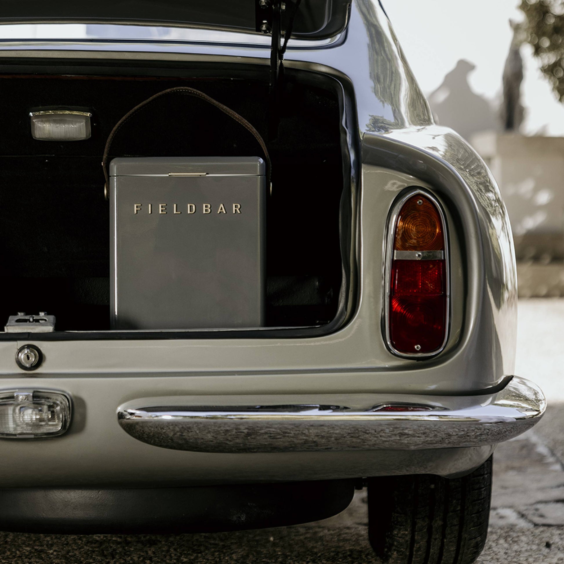 The open trunk of a vintage silver car reveals a gray Fieldbar Drinks Box by Fieldbar, celebrated for its impressive thermal performance. Sunlight creates intriguing shadows on the surrounding wall and ground.