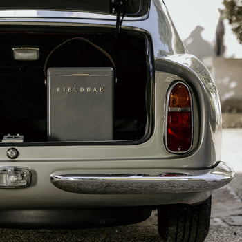 The open trunk of a vintage silver car reveals a gray Fieldbar Drinks Box by Fieldbar, celebrated for its impressive thermal performance. Sunlight creates intriguing shadows on the surrounding wall and ground.
