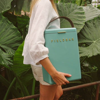 A woman in a white shirt and shorts stands among large green leaves, carrying a teal Fieldbar Drinks Box by Fieldbar with a brown strap over her shoulder.
