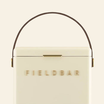 The cream-colored Fieldbar Drinks Box by Fieldbar features gold FIELD BAR lettering, a brown leather top handle, and outstanding thermal performance to keep drinks chilled.