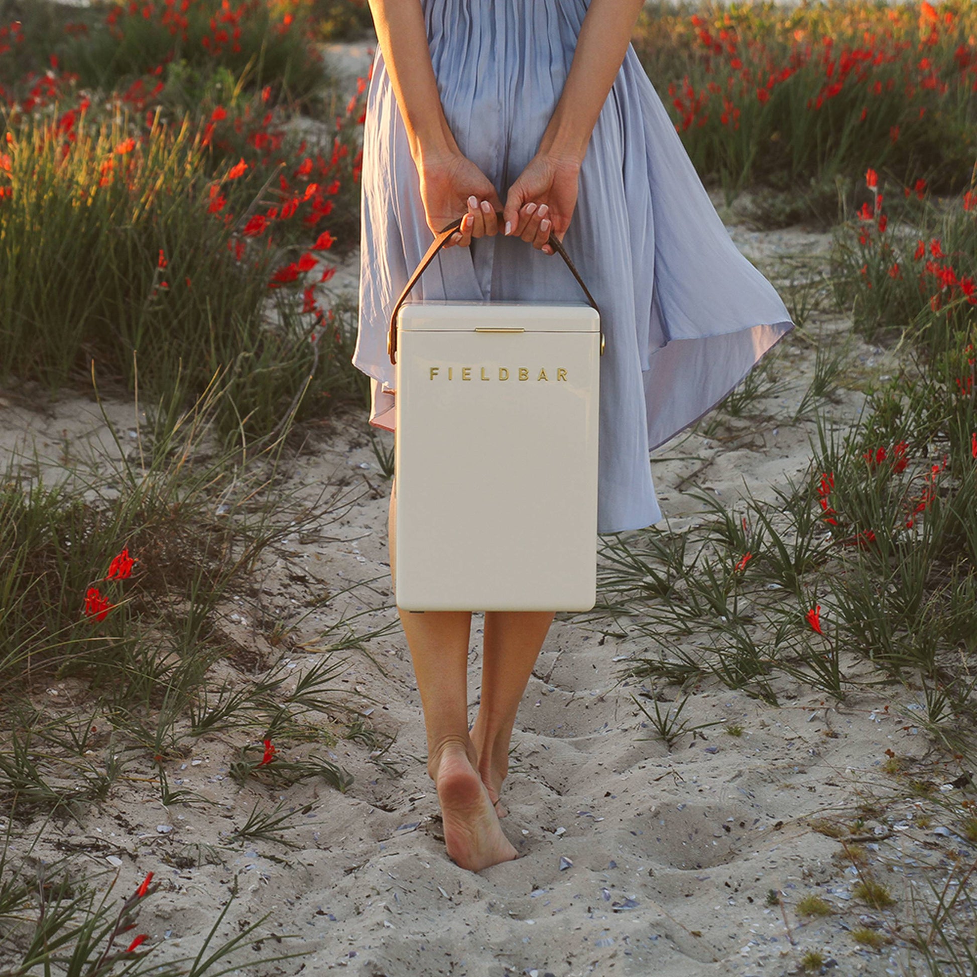 A person in a light blue dress walks barefoot on sandy ground with grass and red flowers, carrying a cream Fieldbar Drinks Box by its handle, ready to enjoy its impressive thermal performance.