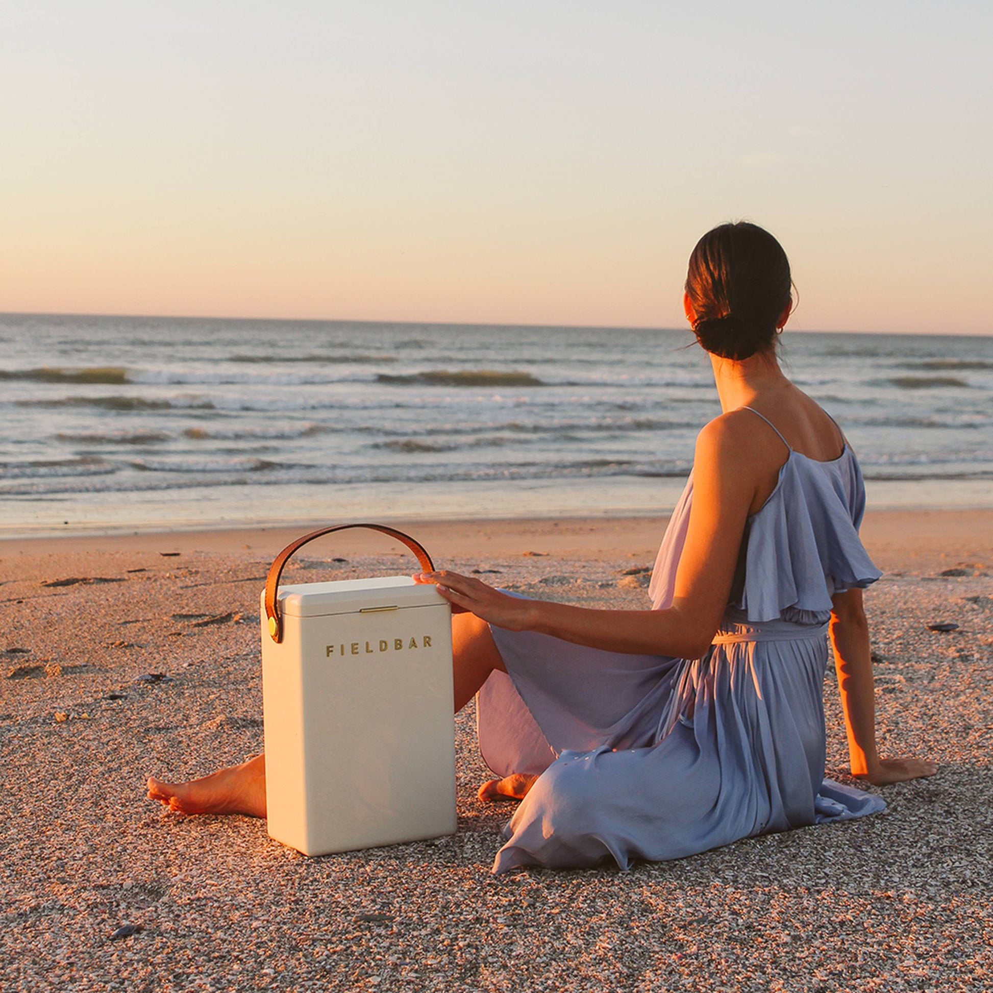 A woman in a light blue dress sits on a sandy beach at sunset, gazing at the sea. Next to her is the Fieldbar Drinks Box by Fieldbar, ensuring her drinks stay perfectly chilled with its dependable thermal performance.