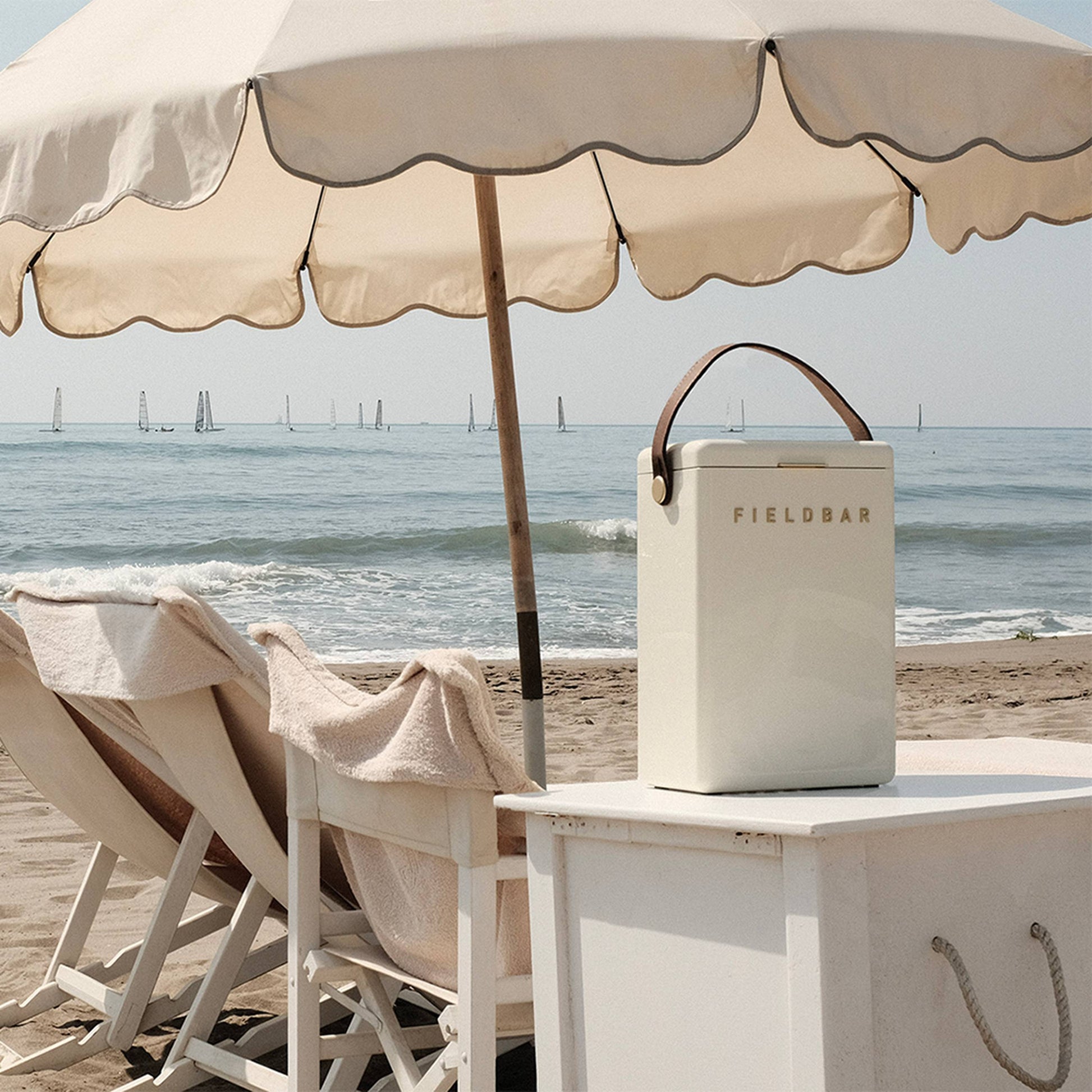 A cream Fieldbar Drinks Box by Fieldbar with a brown handle rests on a white table near lounge chairs and an umbrella on a sandy beach, while sailboats drift across the calm sea in the background.