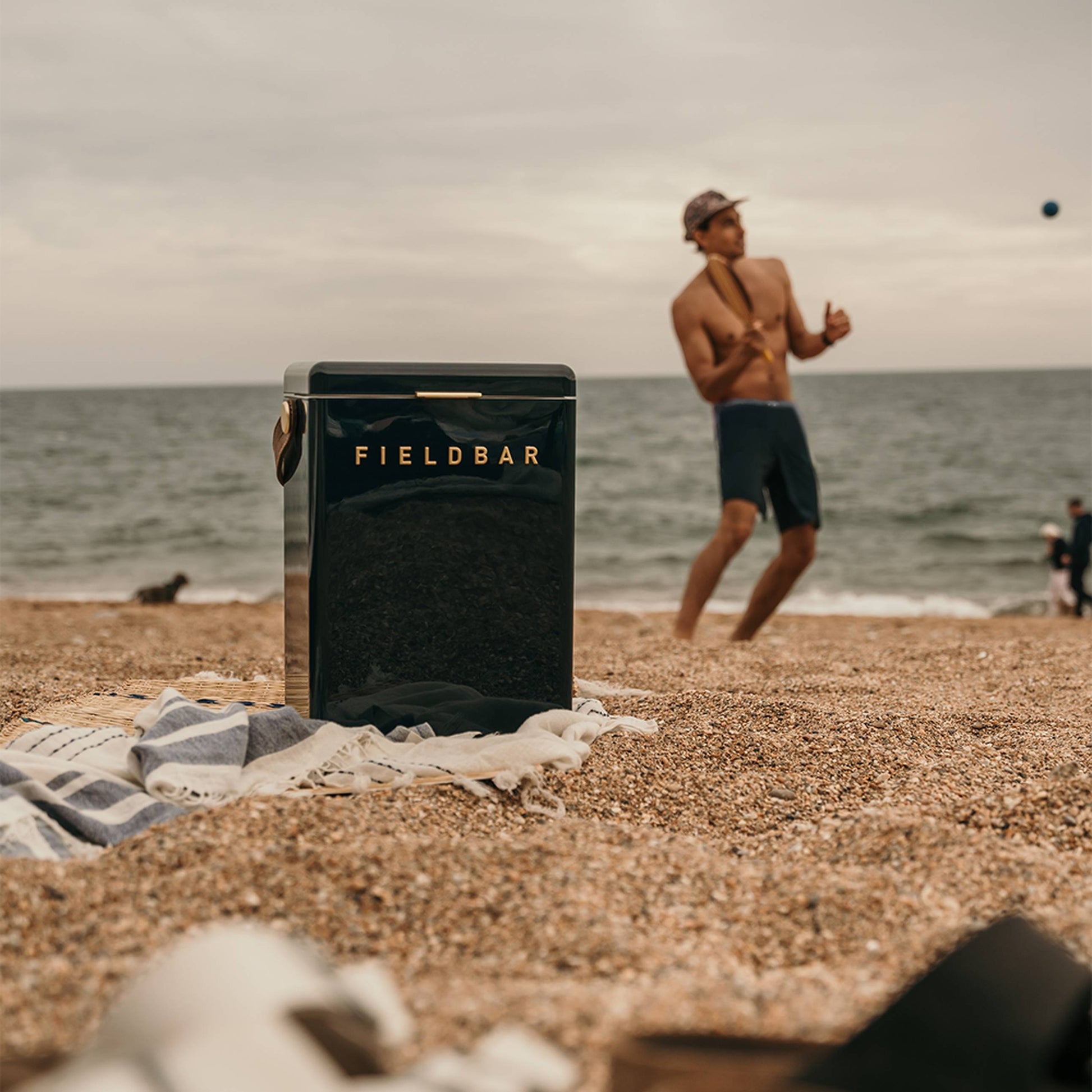 A Fieldbar Drinks Box by Fieldbar rests on a sandy beach next to a towel, as a man in shorts and a cap enjoys the ocean, capturing the ice boxs stylish look and dependable thermal performance in a relaxed summer setting.