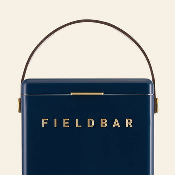 The Fieldbar Drinks Box is a navy blue rectangular cooler with a curved brown handle and gold hardware. FIELD BAR appears in gold on the front. This box provides excellent thermal performance against a light background.