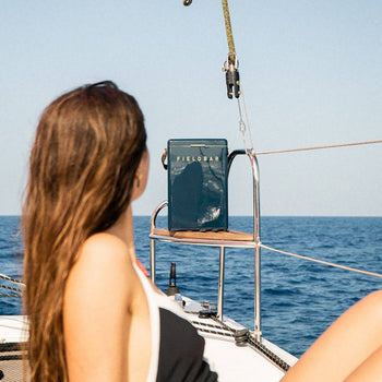A woman in a black swimsuit relaxes on a sailboat, gazing at the Fieldbar Drinks Box by Fieldbar, renowned for its thermal performance, as it keeps drinks cool against the backdrop of the ocean and clear sky.
