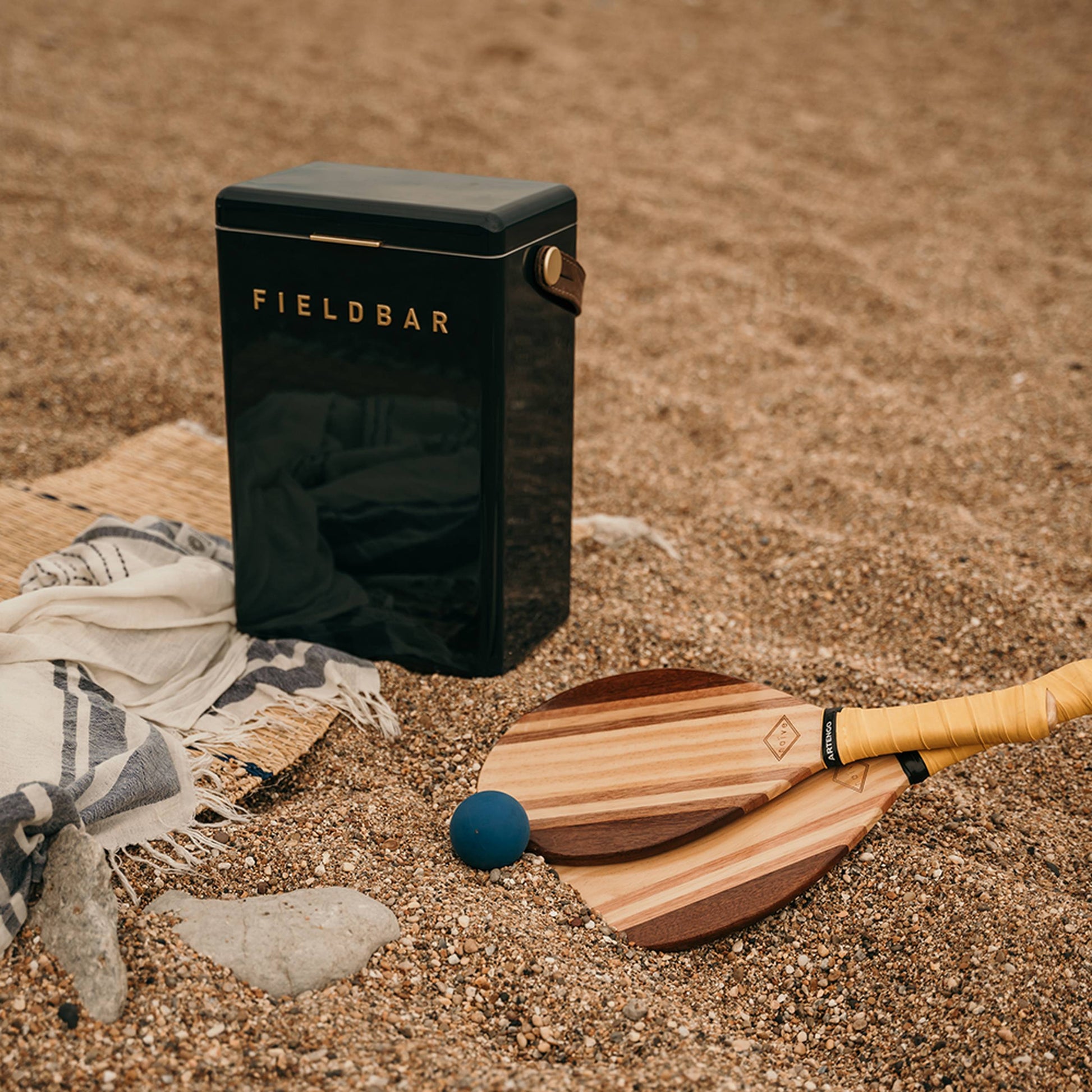 A black Fieldbar Drinks Box from Fieldbar rests on a sandy beach beside a woven mat, striped towel, two wooden bats, and a blue ball, with rocks and pebbles scattered nearby—showcasing its superior thermal performance.