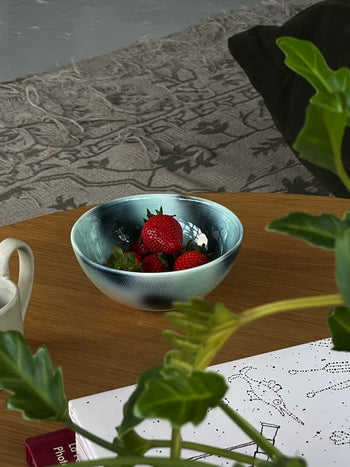 The Frizbee Ceramics BBOL S, a small blue ceramic bowl by Frizbee Ceramics, is filled with strawberries on a wooden table; green leaves, paper, and a patterned rug complete the scene.