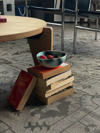 A Frizbee Ceramics BBOL S bowl by Frizbee Ceramics, filled with strawberries, rests on a stack of books by a wooden table atop a patterned carpet, adding artisanal charm to the room with chairs in the background.