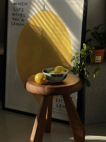The Frizbee Ceramics BBOL S by Frizbee Ceramics sits on a wooden stool with a loose lemon, in front of a vibrant lemon poster and beside a sunlit potted plant.