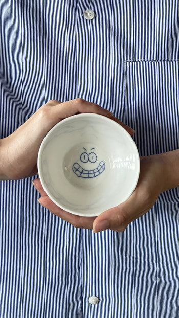 A person in a blue-and-white striped shirt holds the Frizbee Ceramics BBOL S bowl by Frizbee Ceramics, featuring a playful cartoonish smiling face in vivid blue on the inside for an artisanal charm.