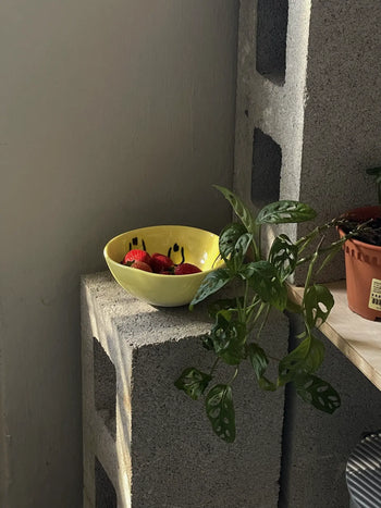 The Frizbee Ceramics BBOL S, a yellow bowl with black smiley faces by Frizbee Ceramics, holds strawberries atop a concrete block beside a potted plant with holey leaves and a shelf in gentle natural light.
