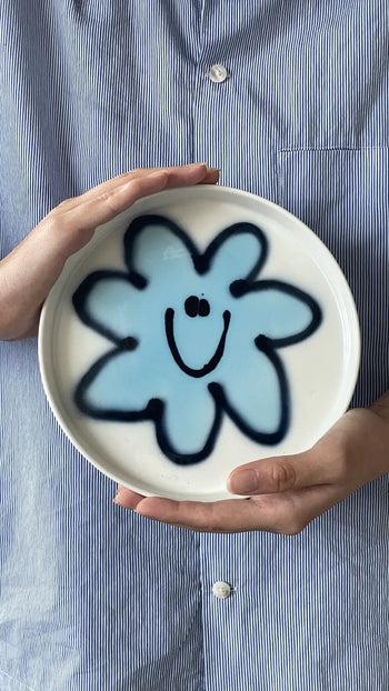 A person in a blue-and-white striped shirt holds the Frizbee Ceramics Baby Plate by Frizbee Ceramics, a round white plate with a blue flower and smiling face at the center, adding artisanal charm to any kitchen collection.