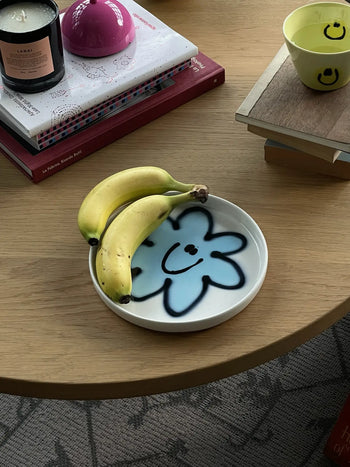Two bananas rest on a Frizbee Ceramics Baby Plate with a blue smiling flower, bringing artisanal charm to a light wooden table set with books, a candle, and a yellow smiley-face cup.