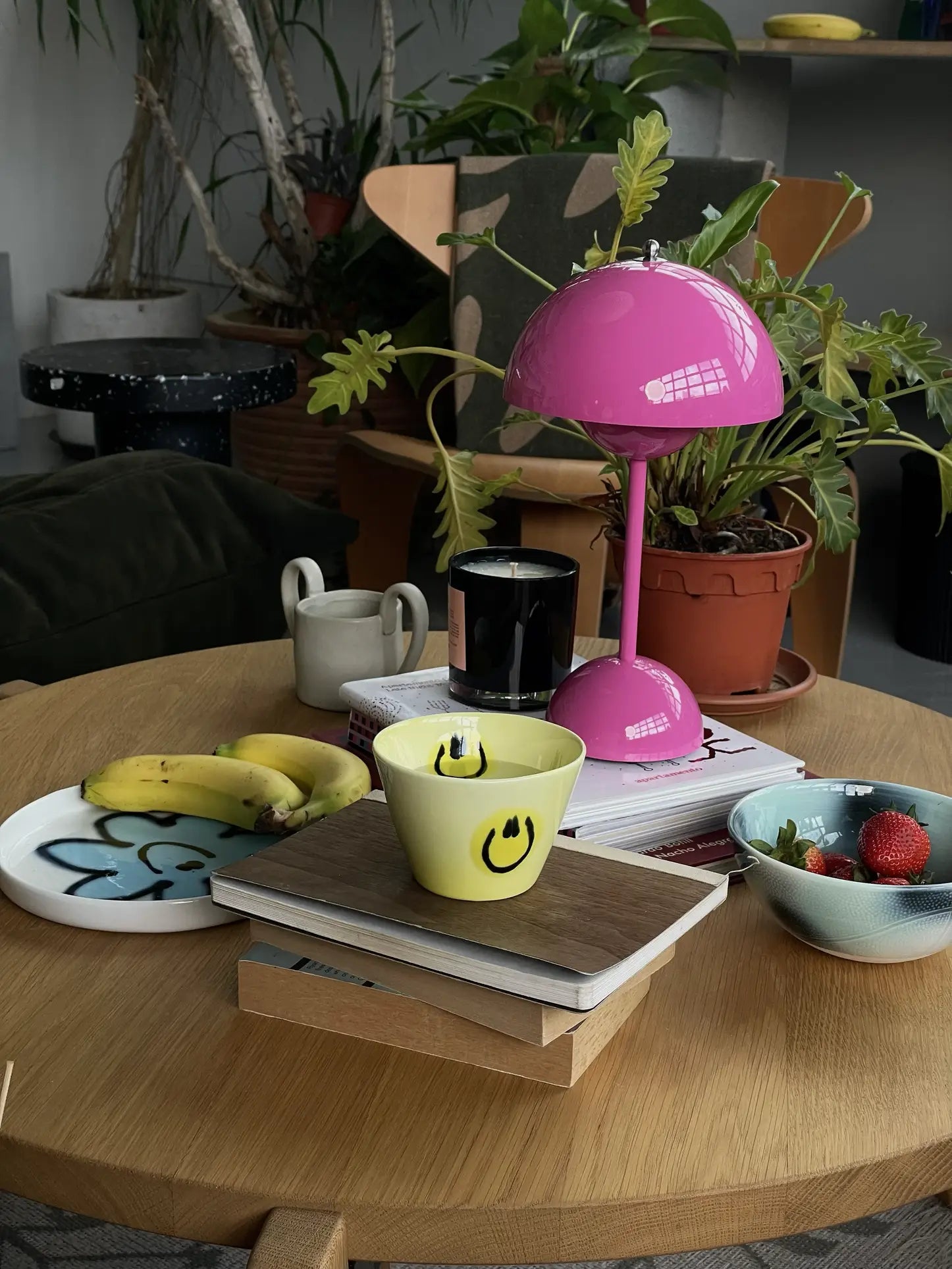 A cozy table scene with a pink lamp, potted plants, yellow smiley cup, bowl of strawberries, bananas on the Frizbee Ceramics Baby Plate by Frizbee Ceramics, a candle, books, and mug—all atop a light wooden table.