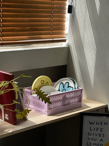 A pink basket with colorful Frizbee Ceramics Baby Plates by Frizbee Ceramics sits on a sunlit wooden desk near brown blinds, alongside a leafy plant and a framed WHEN LIFE GIVES YOU A LEMON sign, adding artisanal charm.